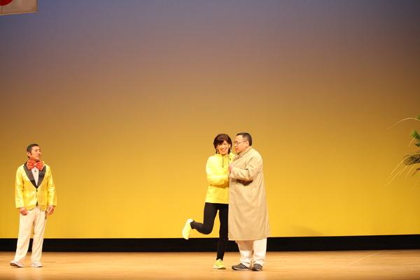 黄色いパーカーを着た女性が、茶色のコートご着た男性の腕に飛びついており、その様子を金色のジャケットに赤い蝶ネクタイの男性が見ている寸劇の場面の写真