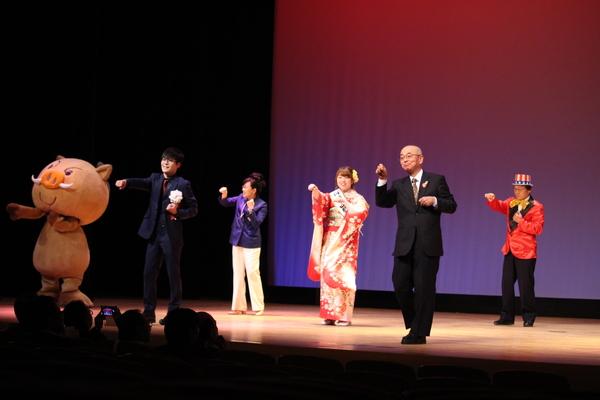 「まるいの」、スーツを着た新成人の男性、紫のジャケットを着た女性、振袖を着た新成人の女性、市長、赤いジャケットに金色の蝶ネクタイを付けた男性が右手に拳をにぎり、突き出して踊っている様な写真