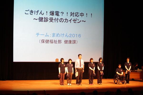 準グランプリ「健康課チーム」7名が壇上に並んでいる写真・改善事例は「ごきげん！爆電？！対応中！！～健診受付のカイゼン～」