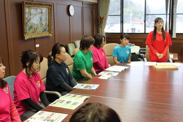 ピンク、緑、水色のシャツを着た複数名の女性が着席し、1名の方が立って話をしている写真