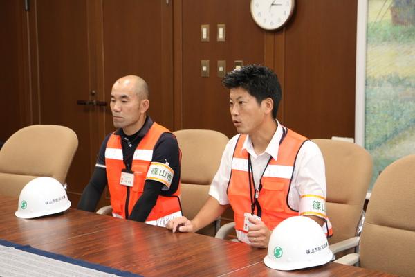 机の上にヘルメットが置いてあり、オレンジ色の安全ベストを着ている市民安全課の小林 智彦主査と農都政策課の森本 良太係長の写真