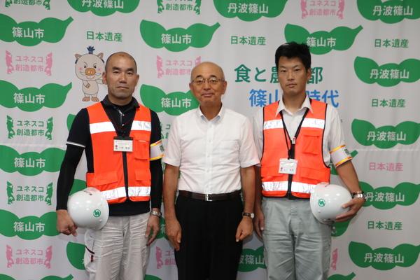 市長を間に挟んで、市民安全課の小林 智彦主査と農都政策課の森本 良太係長の3人が写っている写真