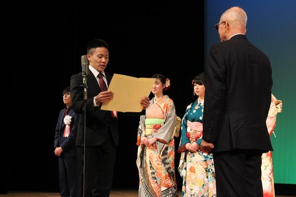 副実行委員長の井谷君が舞台上で、メガネを掛けた黒いスーツの男性の前に立ち、黄色い表紙を両手で持ち、書かれている文章を読み上げている様子の写真