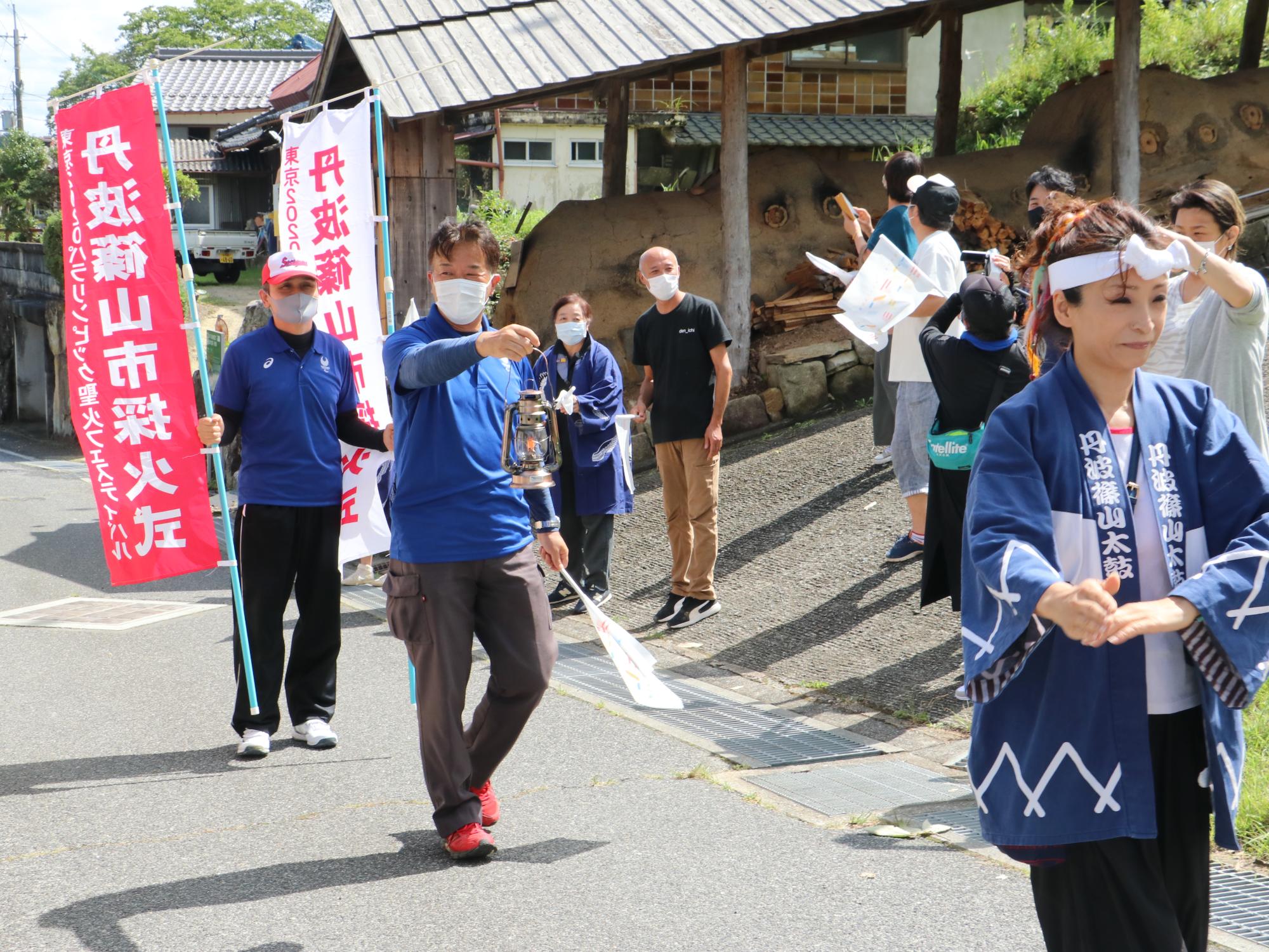 男性が聖火の火をランタンに入れて歩きながら運んでいる。前には法被を着た女性がデカンショを踊りながら歩き、後ろにはのぼり旗を持った男性が歩いている。