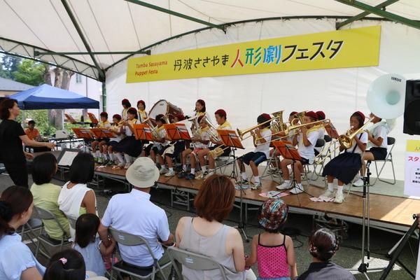 「丹波ささやま人形劇フェスタ」の横断幕の前の舞台で、大芋小学校の金管バンドの演奏を披露しており、お客さんがパイプ椅子に座って聞いている様子の写真