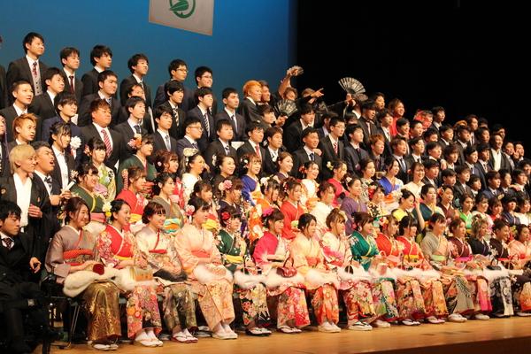 舞台上に出席した新成人が上がっており、前列に振袖を着た新成人が座っており、後方に、スーツを着た新成人が立っており、一番上の中央に袴を着た男性の新成人数名が指差しして写っている集合写真