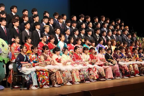 舞台上に出席した新成人が上がっており、前列に振袖を着た新成人が座っており、後方に、スーツを着た新成人が立っており左端に「まめりん」も一緒に写っている集合写真