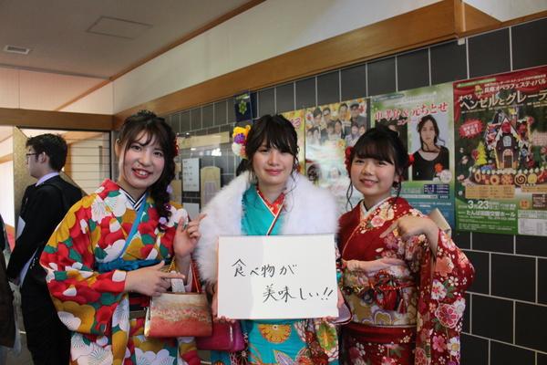 「食べ物が美味しい！！」と書いた画用紙を持って振袖を着た3人の新成人が笑顔で写っている写真