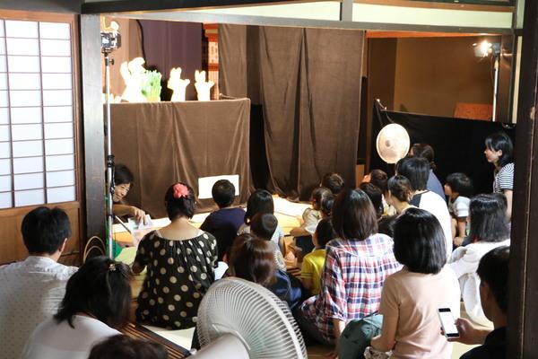 和室の中に茶色い布で覆われた舞台が作られており、人形劇が行われており、沢山の親子連れのお客さんが畳に座って見ている様子を後ろから写している写真