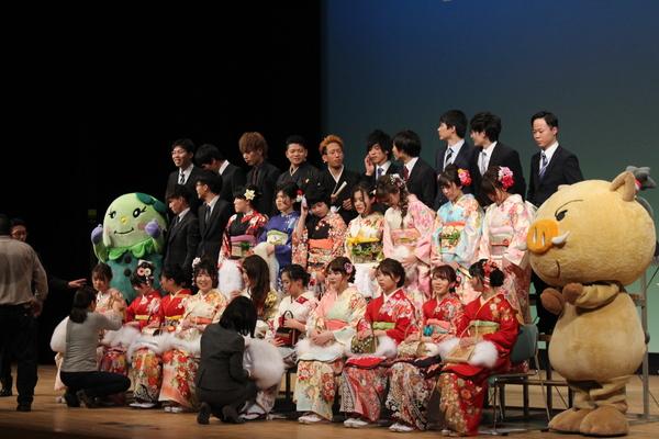 「まめりん」と「まるいの」が新成人の両脇に立っており、集合写真の準備の為新成人達が並んでおり、スーツを着た女性が、新成人の振袖の衣装を直している様子の写真