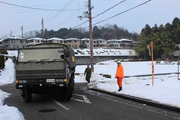 陸上自衛隊のトラックが雪の降り積もっている町の道路を左折して曲がって来ており、道路脇には自衛隊とオレンジ色のジャンパーを着た関係者が立っている様子の写真