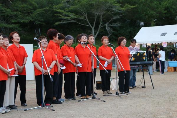 お揃いのオレンジ色のポロシャツを着た女性たちが歌っている写真