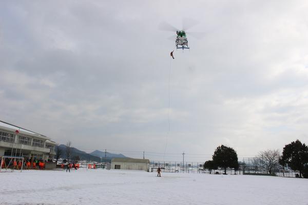 西紀南小学校の運動場の上空にヘリコプターが飛んでおり、ヘリコプターから綱を伝って男性が下りてきており、地上で男性が待っている様子の写真