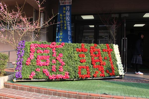 緑色の草や、赤、白、ピンク、紫色の花を使い感謝の文字が書かれた看板が置いてある写真