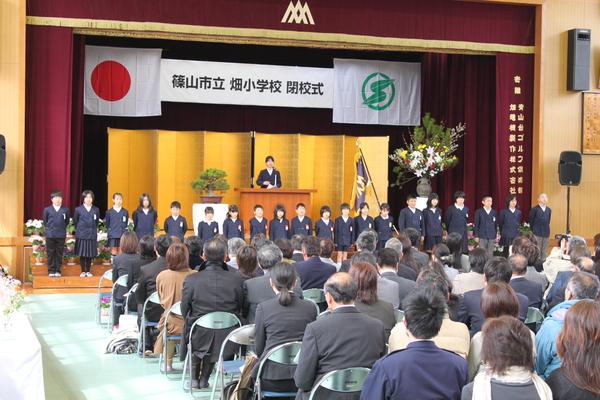 ステージ前に在校生が並び、演説台の前にいる女生徒が保護者に向けて話をしている写真