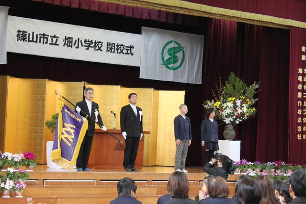 （左から）礼服を着て校旗を持っている男性と直立している男性がおり、その横には畑小学校の代表生徒2名が写っている写真