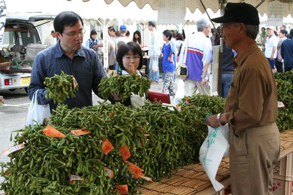 枝豆がたくさん積まれて置いてあり、来場者の男性が枝豆を両手に取って見ている写真