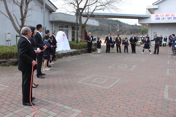 白い布で覆われた物にロープが繋いであり、スーツを着た男性や畑小学校の生徒達がロープを引っ張ろうとしている写真