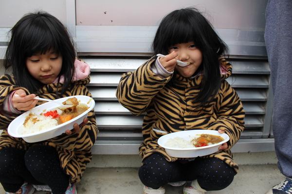 ヒョウ柄のお揃いジャンパーを着た女の子二人が、座ってカレーを食べている様子の写真