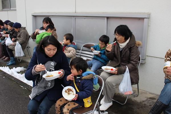 校舎の外に並べられているパイプ椅子に座って、住民のお年寄りや親子がカレーを食べている様子の写真