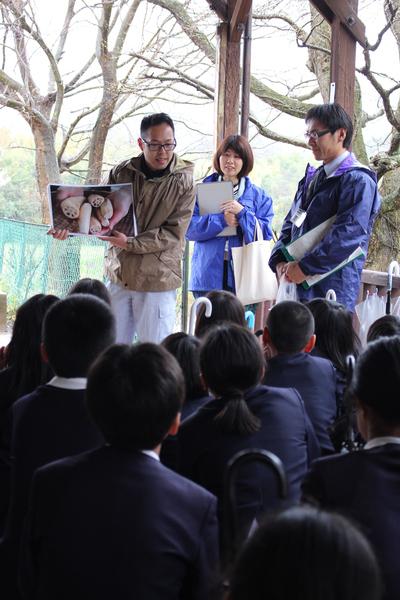 前に先生が2名と農家の方が立っていて、蓮根の写真を持った農家の方がハスの苗の植え付けについて説明しているのを篠山小学校6年生が座って聞いている写真