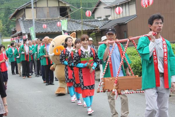 丹波茶まつりの法被を着た男性2人がお賽銭箱を肩に担ぎ、その後方には手に花を持ちながら茶摘みの衣装を着た3名の女性に続き、キャラクター（まるいの）と法被を着た市長や関係者の方々が1列に並んで歩いている写真