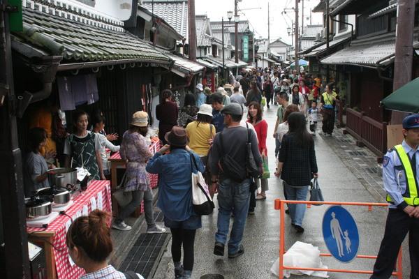 左側に店が並んでいて、手前の店先にはテーブルに鍋が置かれてあり、来場者達が中央の道路を歩いている写真