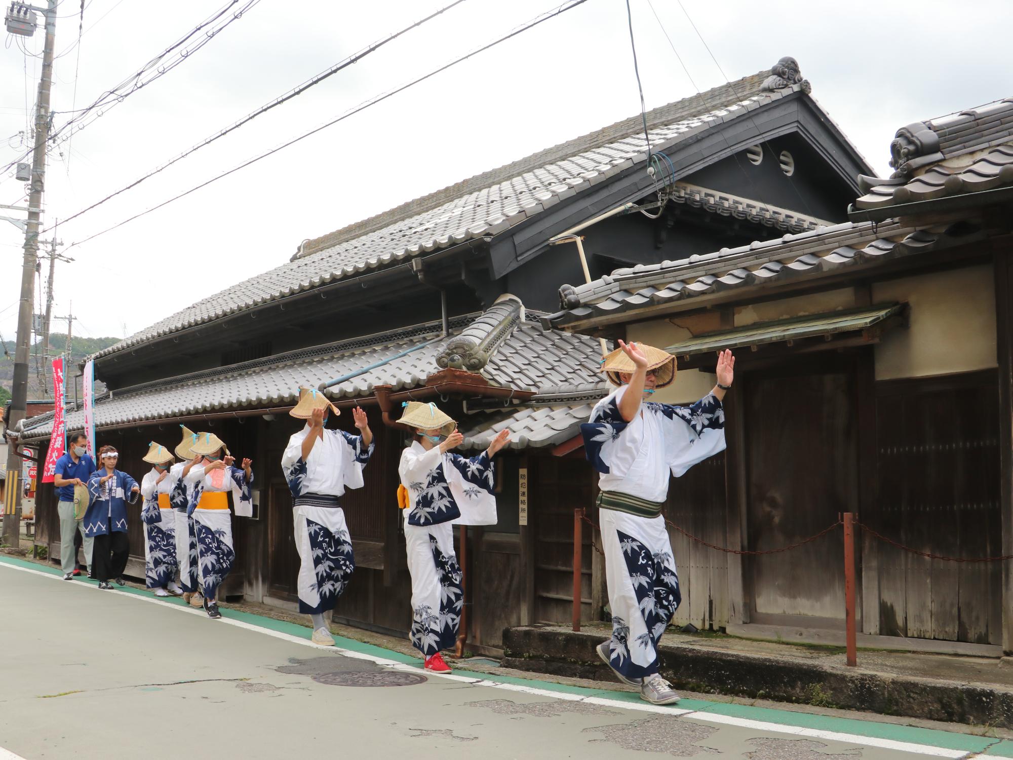 古市の宿場を、編み笠をかぶりデカンショ浴衣を着た6名がデカンショを踊りながら歩いている。列の後方にパラリンピックの聖火がある。