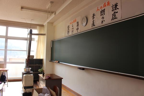 前方に教卓やストーブが置かれ、後ろには黒板があり、その上の壁には謙虚、朗らか、素直、集中、しつけ、整頓と書かれた紙が貼ってある教室内の写真