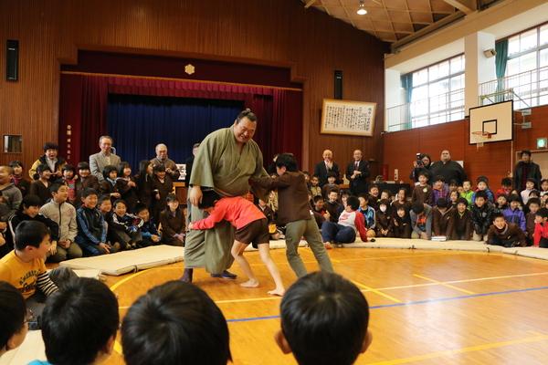 男の子2名が豊ノ島関と相撲を取っており、その周りに集まって応援している小学生と関係者の写真