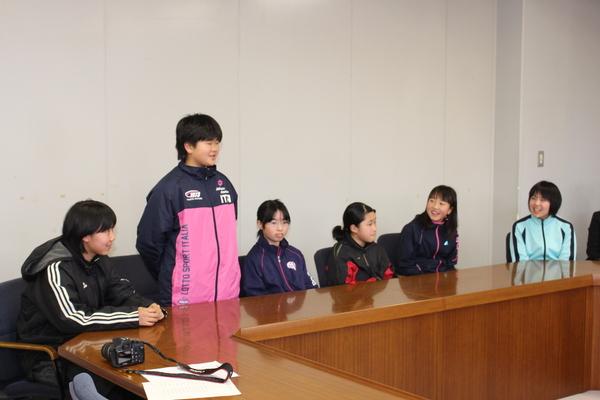 ピンク色ジャンパーを着た女の子が立ち上がって話をしていてその周りに座っている女の子達が笑顔で話を聞いている様子の写真