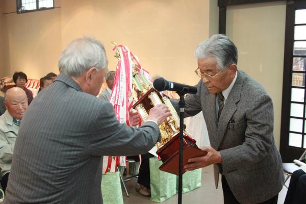 青山賞を獲った酒井 優さんが白髪でメガネをかけた男性から大きなトロフィーを受け取っている写真