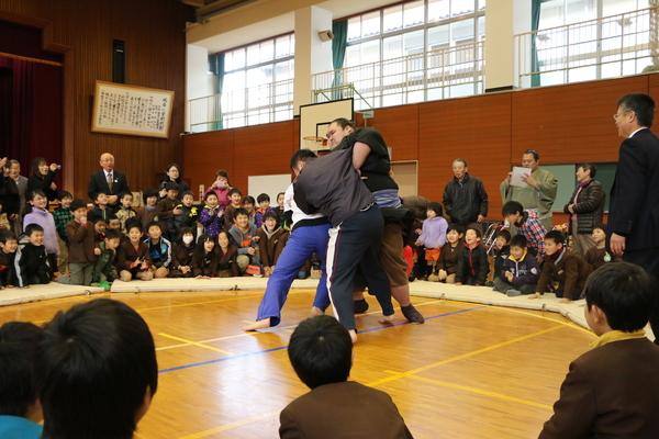 付き人、謙豊と相撲を取る2名の男性と、それを見る小学生と関係者の写真