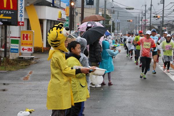 選手が走っている横で虎の帽子をかぶった子と子供達が差し入れをしている写真