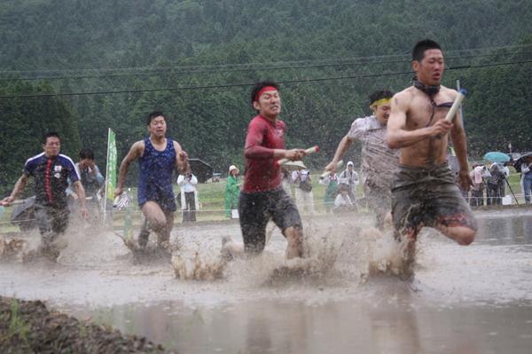 泥の中でバトンを持った男性の方々が水しぶきをあげながらリレーをしている写真