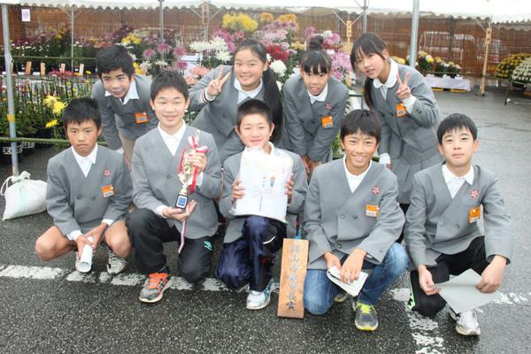 村雲小学校の男女9名が、篠山教育長賞の菊の展示品の前で、トロフィーと記念品を持って嬉しそうに写っている写真