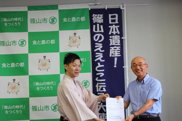 「日本遺産篠山のええとこ伝え大使」と書かれているのぼり旗の前で、袴を着た月亭八斗さんに市長が任命証を渡している写真