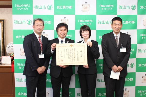 中央の男性と女性が表彰状を手に持って見せ、関係者4名が笑顔で写っている写真