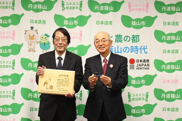 篠山市の文字が入った壁の前で市長と賞状を持った地主 力さんが並んでいる写真