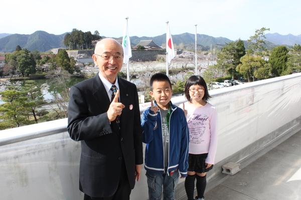 市長室のベランダで男の子と女の子と市長が右手人差し指を立ててポーズをとっている写真