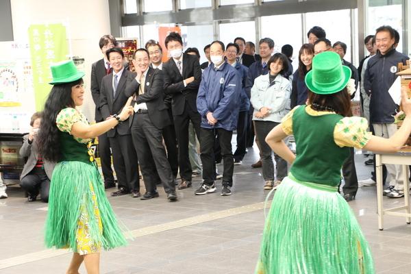緑色の衣装と帽子を被り踊っているココ ポスマンさんと生徒1名を後ろから写し、その踊りを正面から笑顔で見ている人々の写真