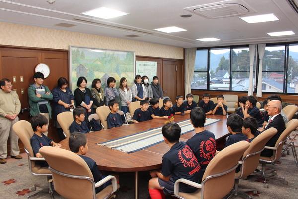 20名ほどのサッカークラブの少年たちが市長と一緒に丸いテーブルの周りに座って話をし保護者の方が立って聞いている写真