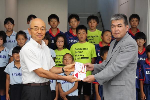 市長と監督が祝い金を持ち、後ろで選手が並んでいる写真