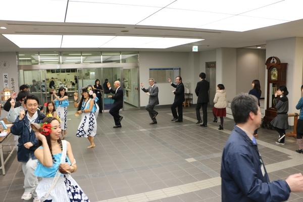ココ ポスマンさんと生徒の皆さんと市の職員の方々がロビーで輪になり、一緒に踊っている写真