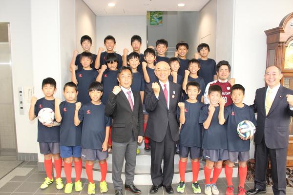 市長と寺本コーチの周りをサッカークラブの少年が囲んで立っていて、全員ガッツポーズをしている写真