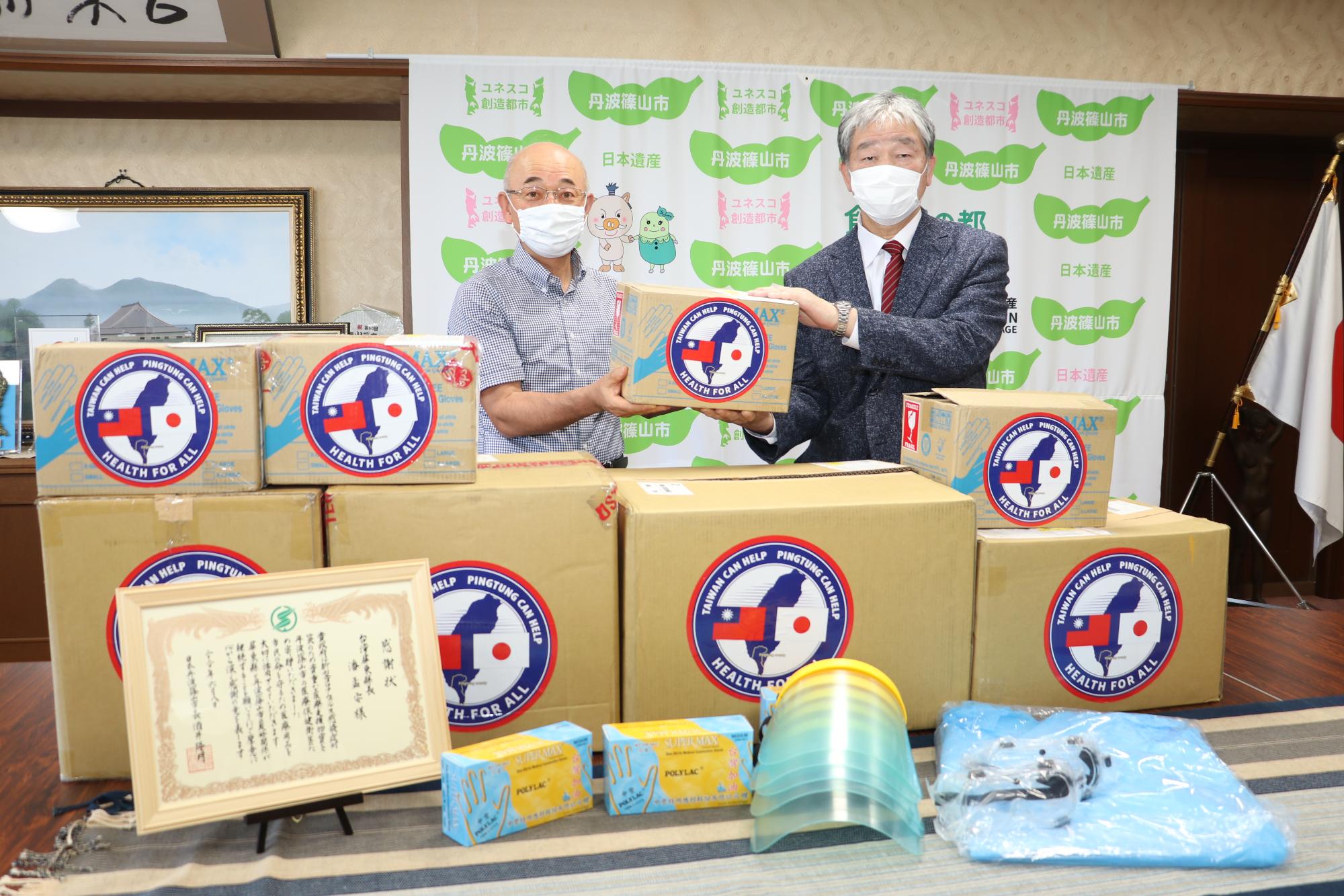 酒井市長と芦田会長が、台湾から届いた支援物資を手にとっている周りには感謝状と手袋、ゴーグル、フェイスシールド、防護服がある