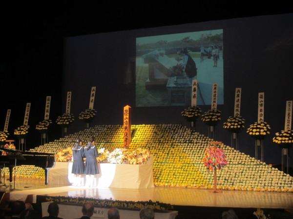 舞台上に作られた菊の花の祭壇の前で、セ－ラー服を着た女子生徒2名が平和宣言文の朗読をしている様子の写真
