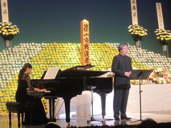 壇上に飾られた菊の花の祭壇の前で、城村 奈都子氏のピアノの伴奏に合わせて、畑儀 文氏が歌を歌っている様子の写真