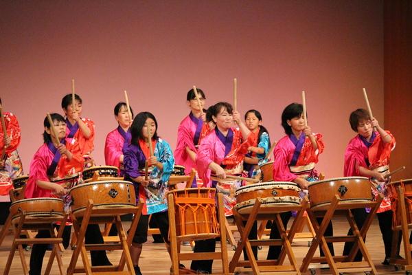 舞台の上で、法被を着て太鼓を演奏する男女の写真