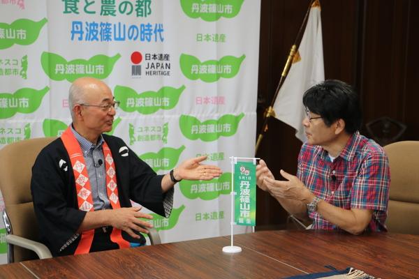 市長と原田さんが、小さい旗を前に座って話をしている写真